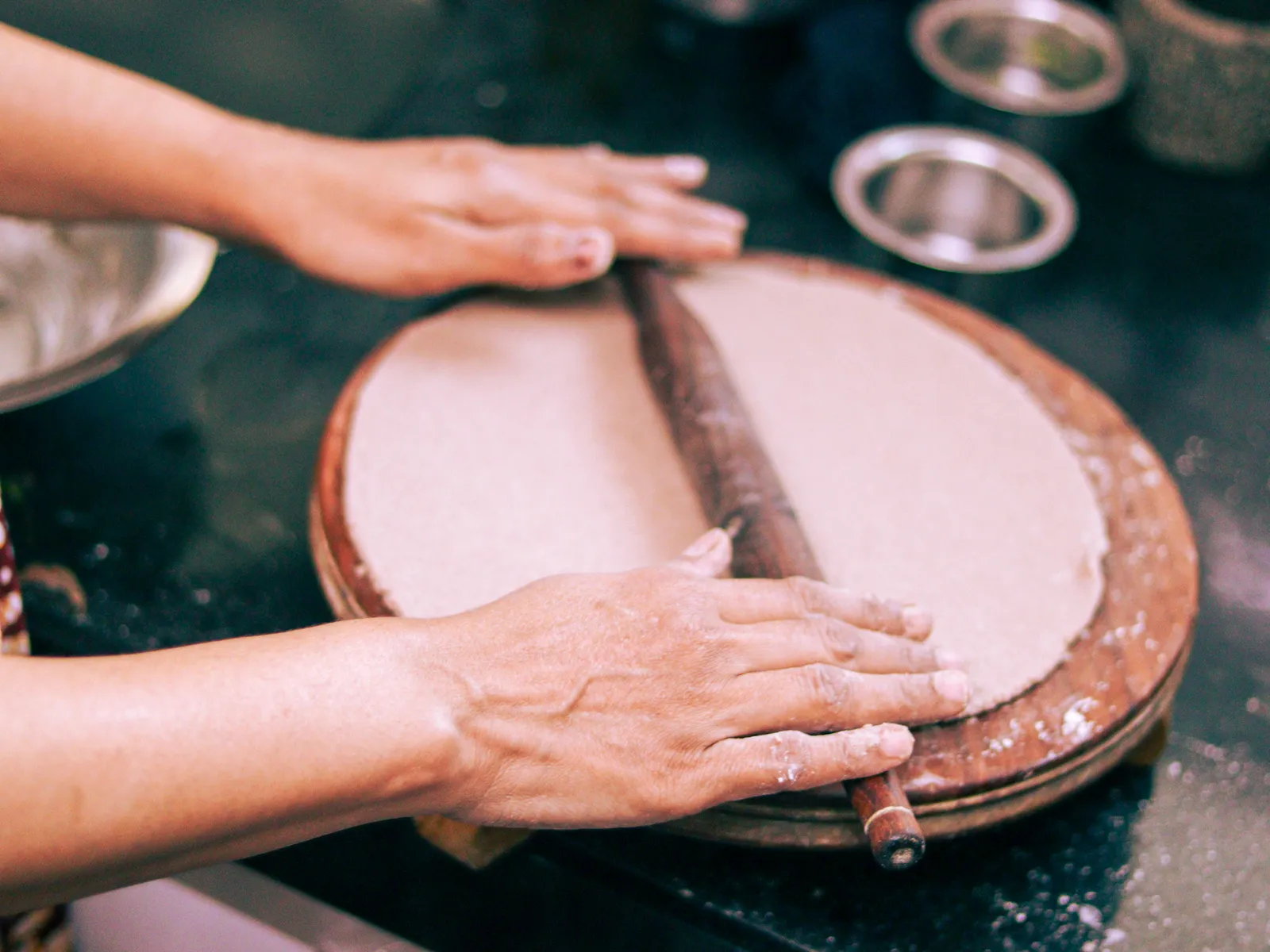 Two hands rolling out a large round of dough on a wooden board, flour dusted on the counter