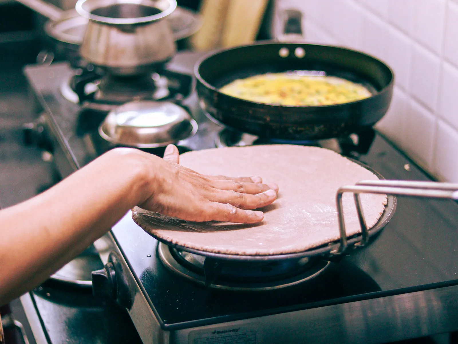 A hand pressing flatbread onto a tawa, an omelette cooking in a pan behind it
