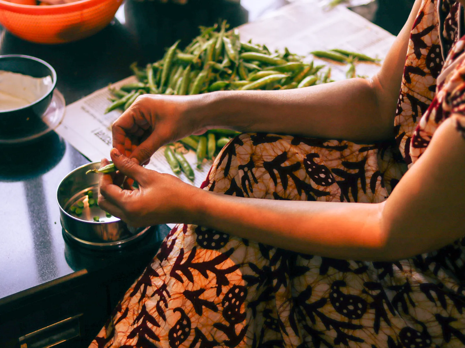 Hands shelling green peas into a small steel bowl, pods heaped on newspaper