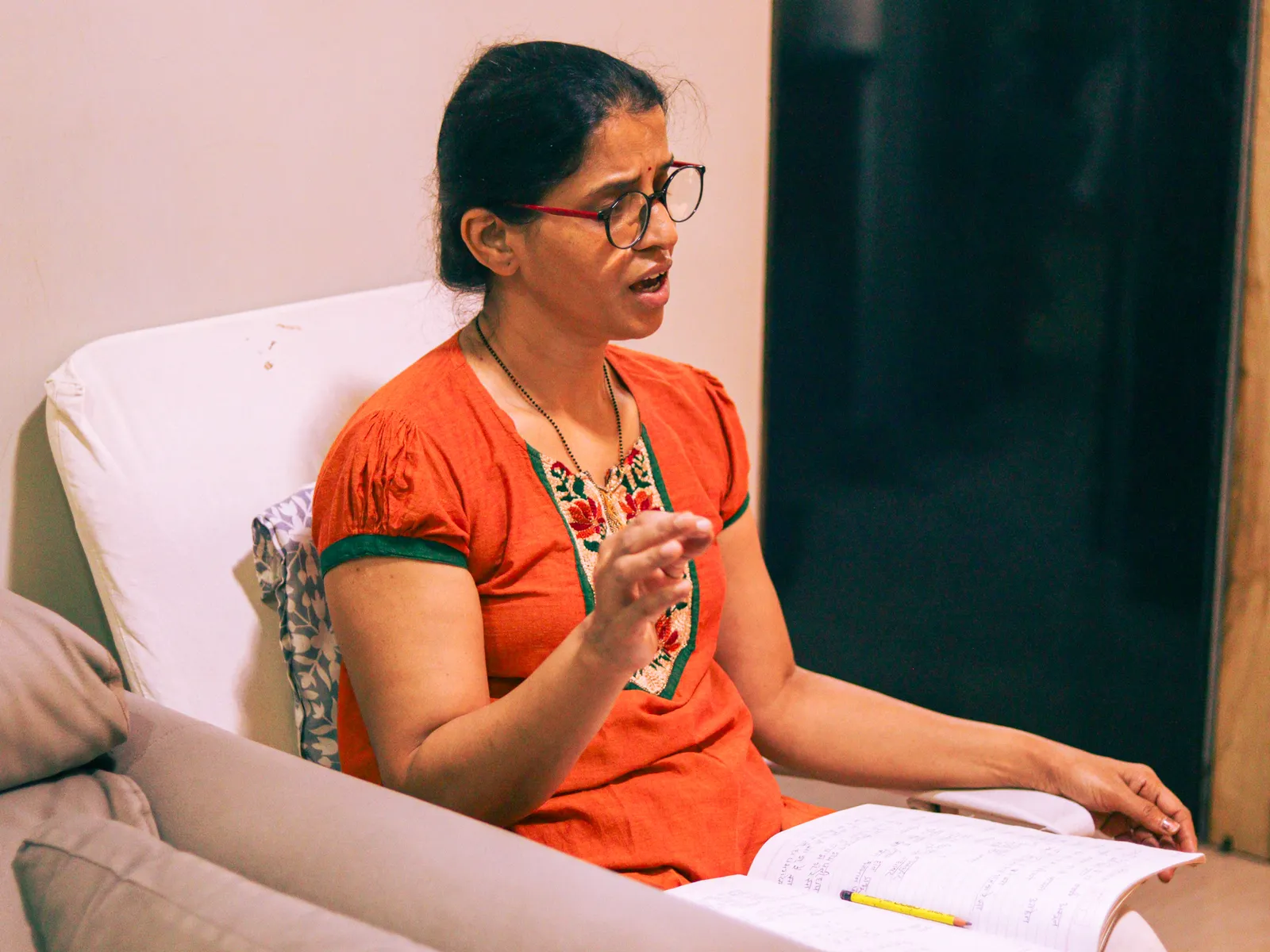 Her practising singing in an orange kurta and glasses, an open notebook on her lap, one hand raised mid phrase